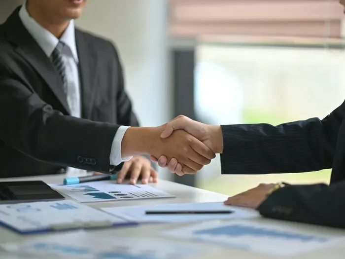 Two business professionals in suits shaking hands across a desk during a meeting with charts and reports